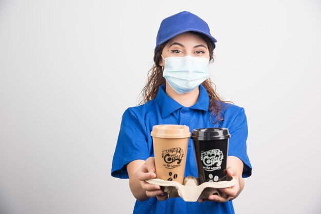 Mulher de uniforme azul com máscara médica segurando duas xícaras de café em branco