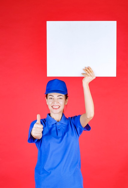 Foto grátis mulher de uniforme azul e boina, segurando uma mesa de informações quadrada branca e mostrando sinal de sucesso com a mão.