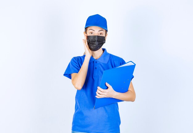 Mulher de uniforme azul e máscara facial preta segurando uma pasta azul e parece confusa e pensativa.