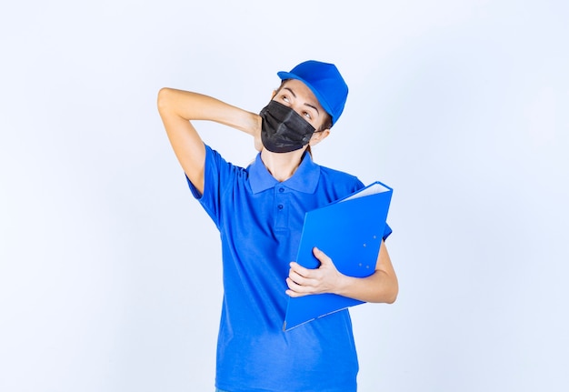 Mulher de uniforme azul e máscara facial preta segurando uma pasta azul e parece confusa e pensativa.