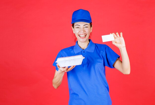 Mulher de uniforme azul segurando uma caixa de plástico branca para levar para entrega e apresentando seu cartão de visita ao cliente.