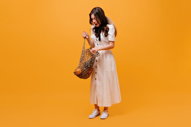 Mulher de vestido branco abre a sacola de compras com frutas e poses em fundo laranja.