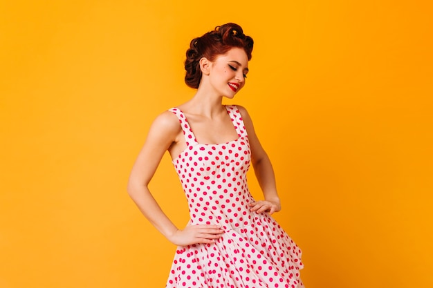 Mulher de vestido de bolinhas, posando com um sorriso. Garota pinup romântica rindo no espaço amarelo.