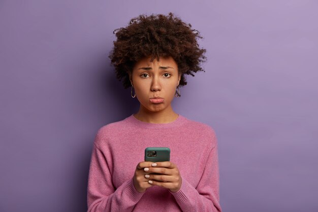 Mulher decepcionada e infeliz com cabelo afro, franze o lábio inferior, segura smartphone, triste por perder chance de uma boa liquidação, chateada por não receber ligação do namorado, posa dentro de casa, vestida casualmente