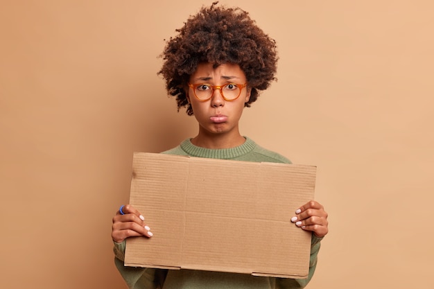 Foto grátis mulher deprimida e triste franze os lábios e olha tristemente para a frente segurando um papelão vazio para seu conteúdo de publicidade e usando óculos óticos isolados sobre uma parede bege