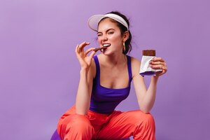 Mulher descolada em roupa esportiva no estilo dos anos 80 morde uma deliciosa barra de chocolate ao leite sentada na parede roxa