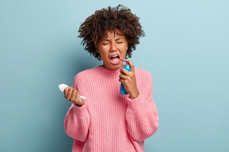 Foto grátis mulher desesperada sofre de dor na garganta, sente-se insalubre e indisposta, espirra spray na boca, segura o lenço de papel, vestida com um macacão enorme, isolada sobre a parede azul. tratamento de gripe ou resfriado