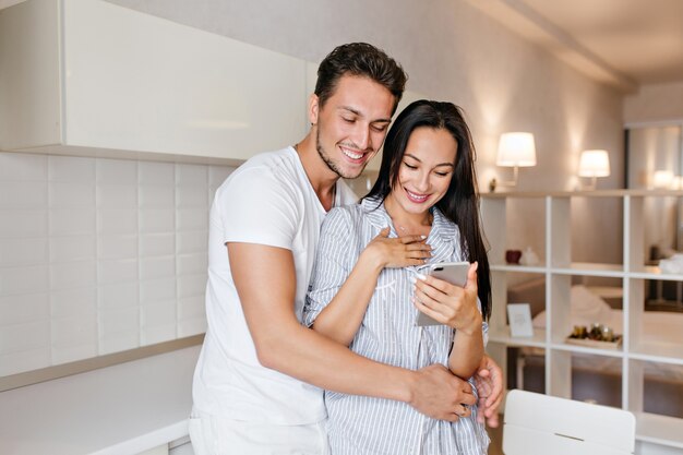 Mulher deslumbrante com sorriso surpreso lendo mensagem de telefone enquanto o marido a abraça