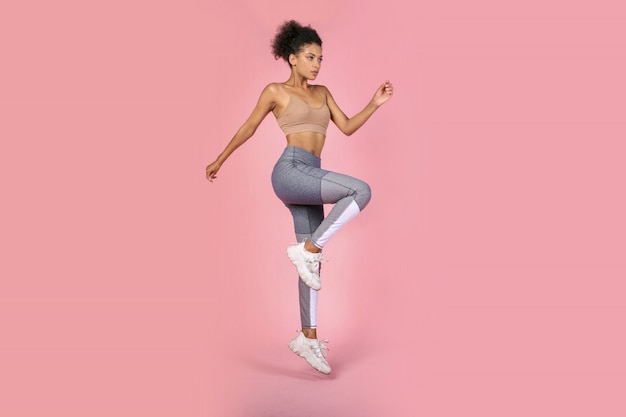 Mulher desportiva praticando agachamento exercícios em estúdio. Mulher africana no sportswear malhando em fundo rosa.
