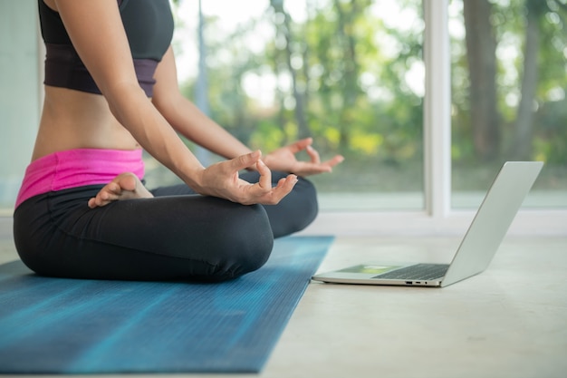 Mulher desportiva praticando ioga, fazendo exercícios ardha padmasana, meditando na posição de lótus, malhando, vestindo roupas esportivas, assistindo a um vídeo tutorial de fitness online no laptop, fazendo exercícios em casa sentado