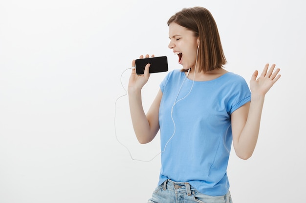 Mulher despreocupada dançando, jogando karaokê no smartphone, cantando no celular e usando fones de ouvido