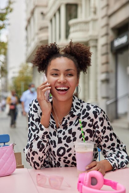 Mulher despreocupada positiva com dois pãezinhos encaracolados fica feliz em ouvir que o melhor amigo faz chamada telefônica bebe milkshake no café de rua gosta de poses de tempo livre contra o fundo da cidade. Estilo de vida urbano