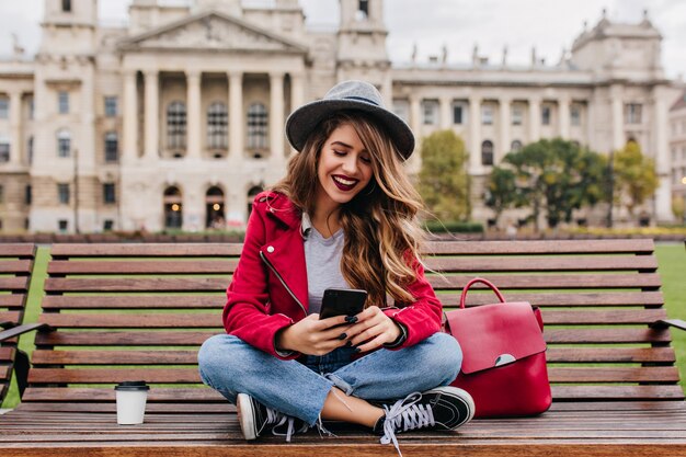 Mulher despreocupada sentada em um banco de madeira enviando mensagens de texto com um sorriso encantador