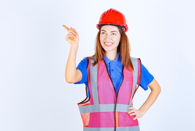 Foto grátis mulher do engenheiro de uniforme e capacete vermelho mostrando o lado esquerdo.