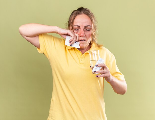 Mulher doente com camisa amarela se sentindo péssima segurando um copo d'água e comprimidos limpando o nariz escorrendo, sofrendo de gripe