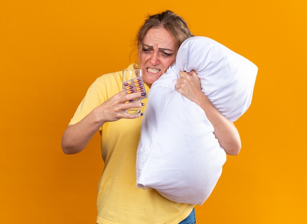 Mulher doente de camisa amarela, com gripe e resfriado, sentindo-se mal, abraçando o travesseiro, segurando comprimidos e um copo d'água, parecendo irritada e desapontada em pé sobre a parede laranja