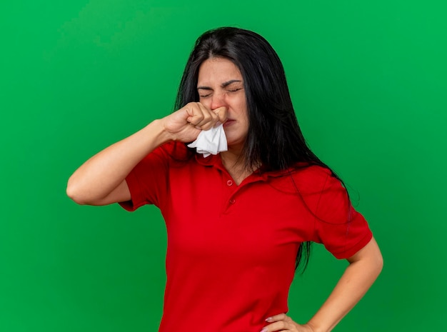 Mulher doente e carrancuda segurando um guardanapo, colocando a mão na cintura, limpando o nariz com os olhos fechados, isolada na parede verde