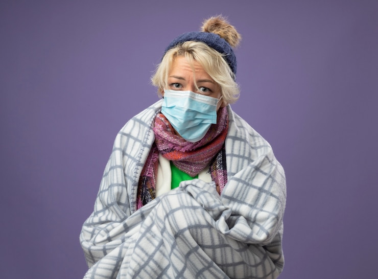Foto grátis mulher doente e insalubre com cabelo curto, chapéu quente e lenço usando máscara facial protetora envolta em cobertor, sentindo-se mal olhando para a câmera infeliz sobre o fundo roxo