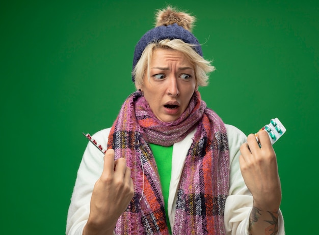 Foto grátis mulher doente e insalubre com cabelo curto em um cachecol quente e um chapéu se sentindo mal segurando bolhas com comprimidos, olhando para a câmera, preocupada e confusa, tentando fazer uma escolha em pé sobre um fundo verde