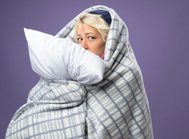 Foto grátis mulher doente e insalubre com cabelo curto em um chapéu quente enrolado em um cobertor segurando um travesseiro, sentindo-se mal e com uma expressão triste preocupada com a parede roxa