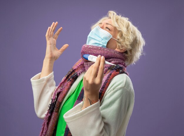 Mulher doente e insalubre com cabelo curto em um lenço quente e máscara protetora facial segurando o termômetro feliz e animada de pé sobre fundo roxo