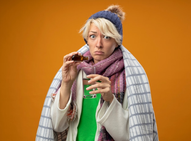 Foto grátis mulher doente e insalubre com cabelo curto em um lenço quente e um chapéu embrulhado em um cobertor pingando remédio que cai em um copo olhando para a câmera confusa em pé sobre um fundo laranja