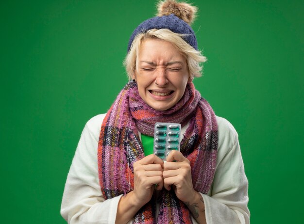 Mulher doente e insalubre com cabelo curto em um lenço quente e um chapéu se sentindo mal segurando uma bolha com comprimidos com expressão irritada no rosto em pé sobre um fundo verde