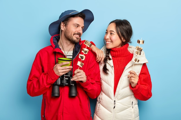 Mulher e homem alegres acamparam juntos, seguram saboroso marshmallow feito na fogueira, olham um para o outro com sorriso, passam o tempo de lazer na natureza selvagem carregando binóculos