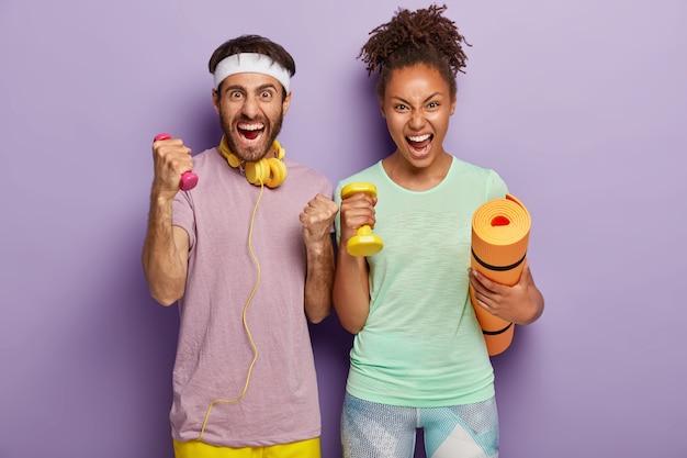 Foto grátis mulher e homem de raça mista emocional gritam alto, seguram karemat e pesos, treinam com o treinador, gritam de desespero, cansados de treinar, isolados na parede roxa. pessoas, esporte, estilo de vida