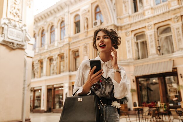 Mulher elegante com blusa clara com laço, segurando a bolsa escura e o telefone na cidade. Mulher de cabelo ondulado com lábios brilhantes desvia o olhar para a rua.