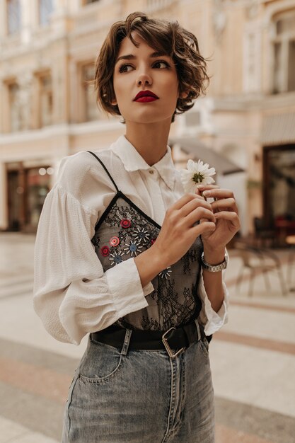 Mulher elegante com cabelo curto em jeans com cinto segurando flores na rua. Mulher de blusa branca com renda preta, posando na cidade.