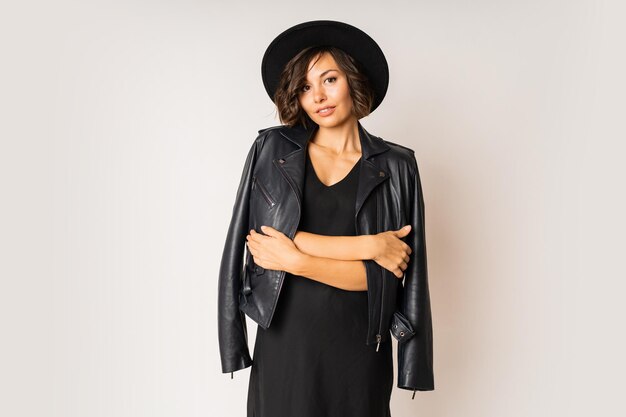 Mulher elegante com chapéu preto, vestido de noite e jaqueta de couro, posando em branco.