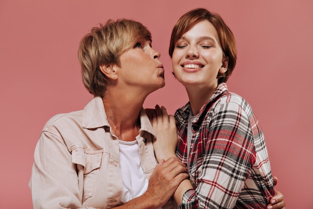 Mulher elegante com penteado curto loiro em roupas bege, beijando na bochecha garota sorridente com roupa xadrez em fundo rosa.