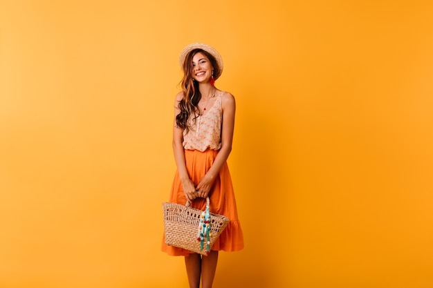 Mulher elegante com roupa de verão se preparando para as férias. Romântica garota ruiva com chapéu de palha, posando em laranja com saco.