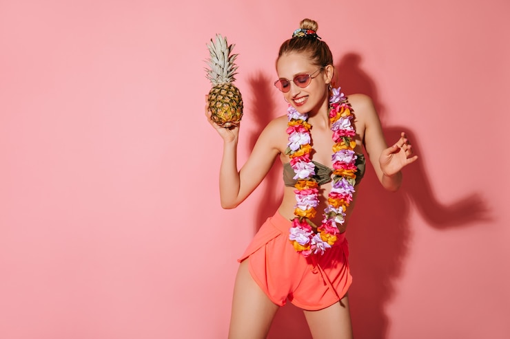 Foto grátis mulher elegante com sorriso encantador em óculos de sol, shorts, maiô e colar de flores brilhantes dançando com abacaxi na parede rosa