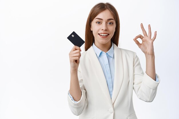 Mulher elegante de terno de negócios mostra cartão de crédito de plástico e sinal de tudo bem sob controle fica contra fundo branco