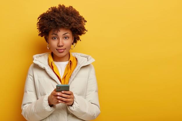 Mulher elegante e atraente com casaco branco quente, olha diretamente para a câmera, usa telefone celular moderno para bater papo online, tem corte de cabelo encaracolado, isolado sobre fundo amarelo. pessoas e tecnologia moderna