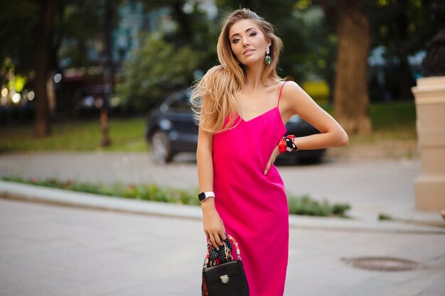 Mulher elegante e atraente usando um vestido rosa sexy de verão andando na rua segurando uma bolsa