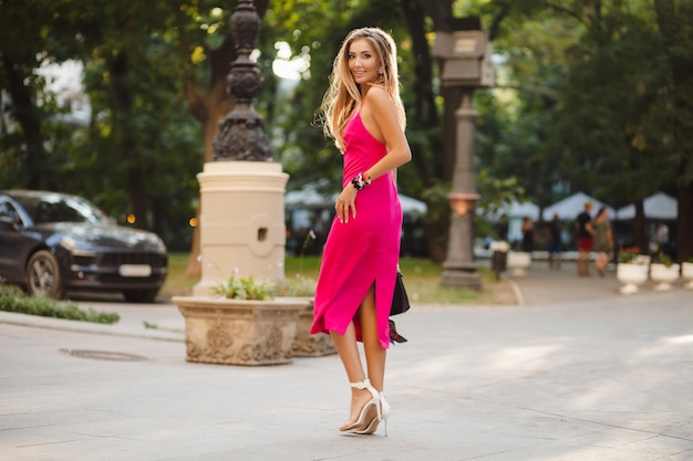 Mulher elegante e atraente usando um vestido rosa sexy de verão andando na rua segurando uma bolsa