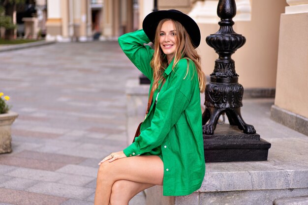 Mulher elegante e elegante posando no centro da cidade europeia, vestindo terno verde de linho brilhante e chapéu preto, estilo de férias de verão.