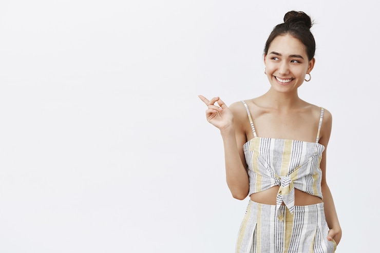 Foto grátis mulher elegante e feliz com pele bronzeada e verruga sob os lábios, sorrindo alegremente enquanto aponta e olha para a esquerda, aproveitando a noite formal dedicada ao seu aniversário