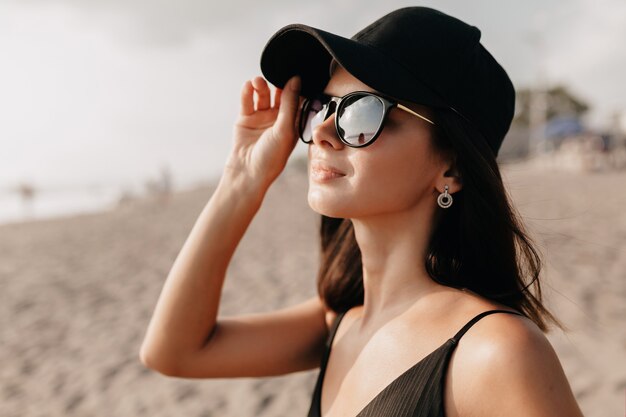 Mulher elegante e moderna em roupas da moda olhando para o oceano com um sorriso feliz usando boné e óculos e aproveita os dias quentes de verão. Jovem modelo feminina, branca