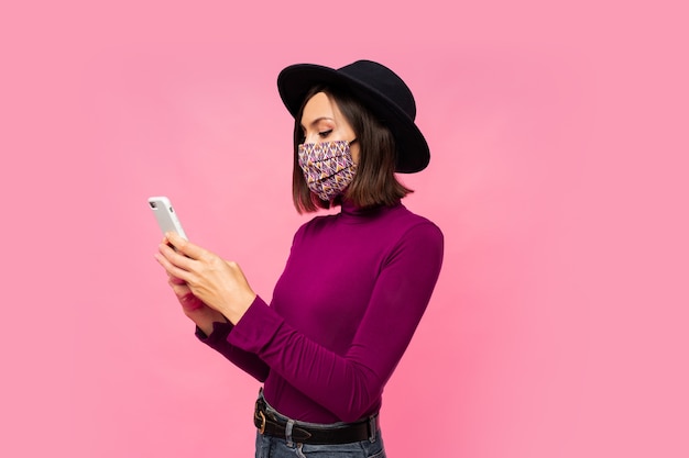 Foto grátis mulher elegante em máscara protetora e chapéu preto, usando telefone celular, em pé.