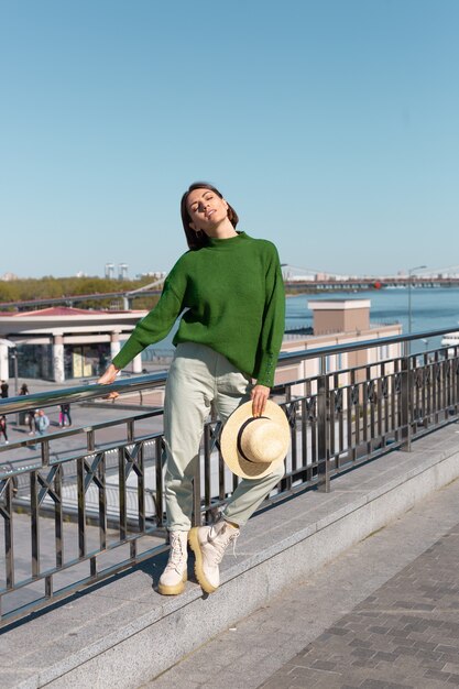 Mulher elegante em suéter verde casual e chapéu ao ar livre na ponte com vista para o rio aproveita um dia ensolarado de verão