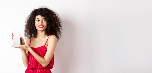 Mulher elegante em vestido vermelho e maquiagem mostrando a tela do smartphone em branco e sorrindo em pé sobre o branco