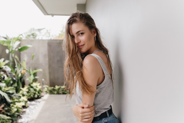 Mulher elegante magro olhando por cima do ombro durante o fim de semana. Retrato ao ar livre do modelo feminino bonito com penteado longo em pé perto de casa.