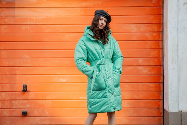 Foto grátis mulher elegante posando com casaco de baiacu de tendência da moda outono inverno e boina de chapéu contra uma parede laranja na rua