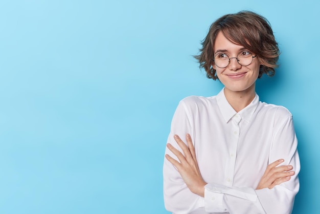 Mulher elegante satisfeita se sente satisfeita mantém os braços cruzados considera sugestão desvia o olhar com expressão feliz usa óculos redondos camisa branca formal isolada sobre o espaço de cópia de fundo azul