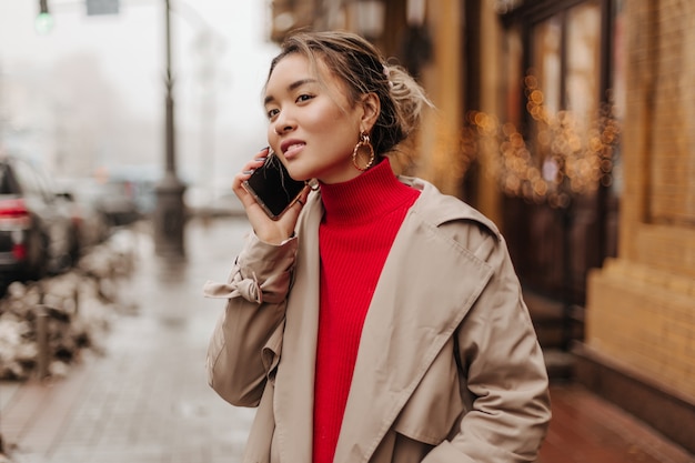 Mulher elegante usando sobretudo leve e suéter brilhante falando ao telefone com ótimo humor e caminhando pela cidade