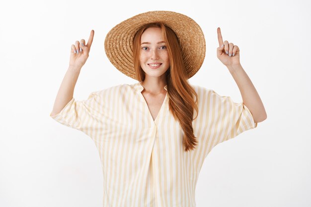 Mulher em pé com uma blusa amarela da moda e um chapéu de palha de verão, levantando os braços, apontando para cima e sorrindo alegremente sobre a parede cinza